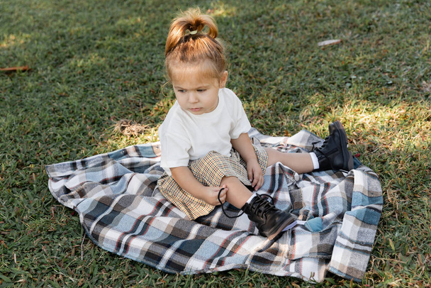 baby girl in checkered skirt and boots sitting on blanket during picnic  - Photo, Image