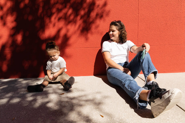 fröhliche Mutter in Jeans schaut Kleinkind-Tochter an, während sie in der Nähe der roten Wand sitzt  - Foto, Bild