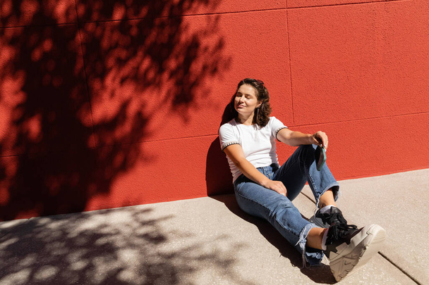 cheerful woman in white t-shirt and blue jeans sitting near building with red wall  - Zdjęcie, obraz