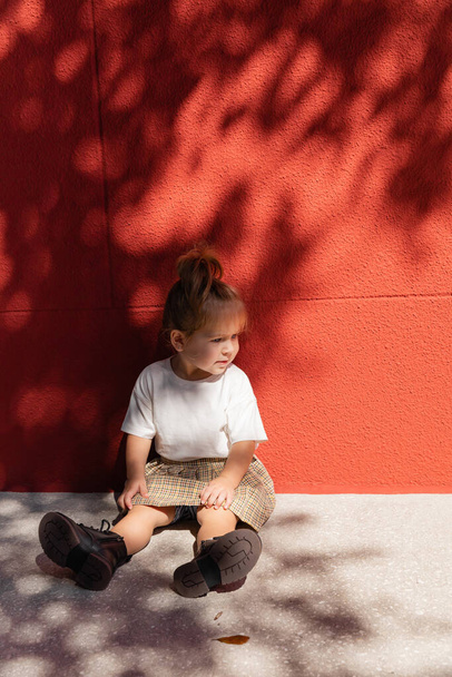 toddler girl in checkered skirt and white t-shirt sitting near building with red wall  - Photo, Image