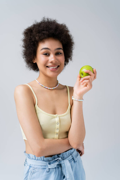 Positive african american model in top holding apple isolated on grey  - Fotó, kép