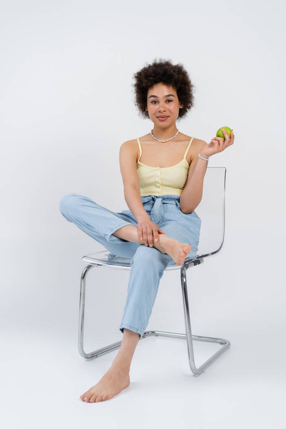 Femme afro-américaine pieds nus tenant pomme fraîche tout en étant assis sur une chaise sur fond gris  - Photo, image