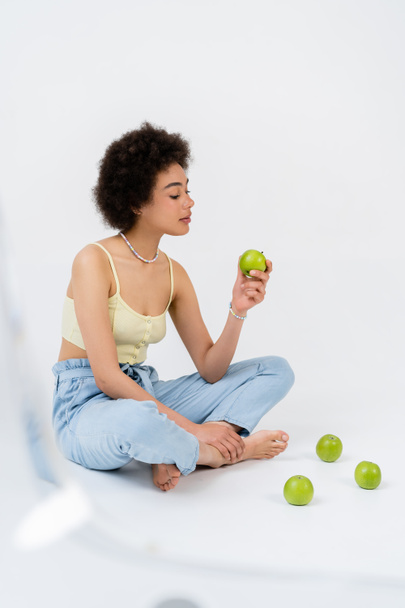 Young african american woman in top holding green apple while sitting on grey background  - Valokuva, kuva
