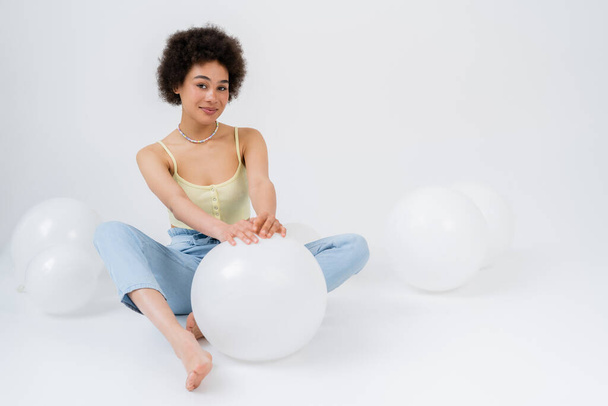 Barefoot african american woman looking at camera near balloons on grey background  - Foto, imagen