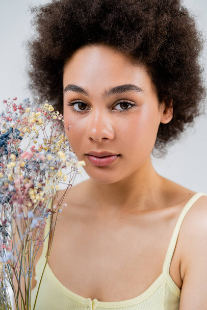Portrait of african american woman with natural makeup looking at camera near baby breath flowers isolated on grey  - Foto, afbeelding