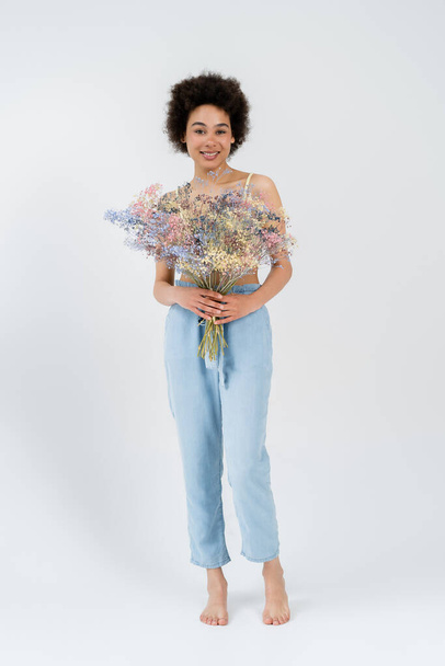 Full length of barefoot african american woman holding baby breath flowers on grey background  - Foto, Imagem
