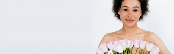 Pequeña mujer afroamericana con hombros desnudos posando con tulipanes aislados en gris, pancarta  - Foto, imagen