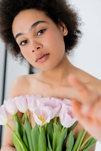 African american woman with naked shoulders holding tulips and pointing with finger on grey background  - Valokuva, kuva