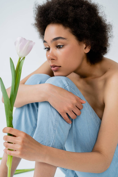 Curly african american model with naked shoulders holding tulip while sitting on grey background  - Φωτογραφία, εικόνα