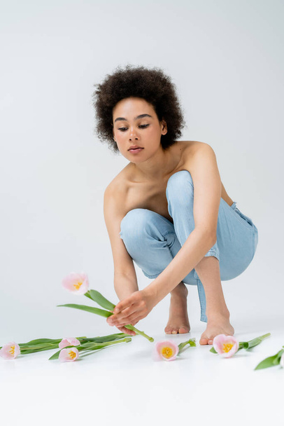 Descalza mujer afroamericana con hombros desnudos tomando tulipán sobre fondo gris  - Foto, Imagen
