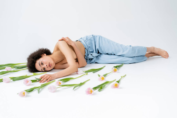 Американка без рубашки и босиком трогает цветок тюльпана, лежа на сером фоне.  - Фото, изображение