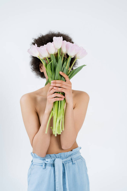 Afroamerykanka bez koszuli trzymająca tulipany w pobliżu twarzy odizolowana na szaro  - Zdjęcie, obraz