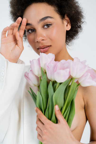Portrait of african american model in jacket holding bouquet isolated on grey  - 写真・画像
