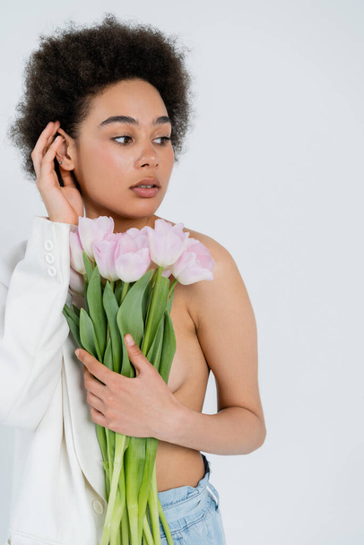 Stylish african american woman in jacket with naked shoulder holding flowers isolated on grey  - Photo, Image