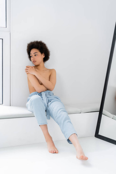 Shirtless and barefoot african american woman siting on windowsill on grey background  - Photo, Image