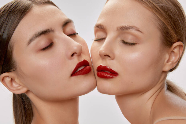 Un toque ligero. Dos sensuales mujeres jóvenes hermosas con maquillaje desnudo y lápiz labial rojo posando con los ojos cerrados. Concepto de amor, emociones, belleza, visión, salud y moda - Foto, imagen