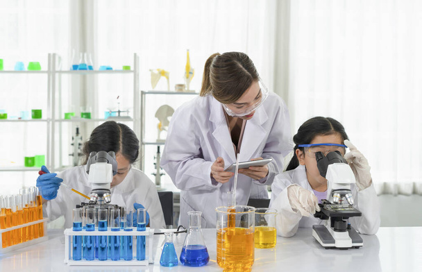 naispuolinen tiedemies mentorina opettaa opiskelijoille aasialaisia lapsia tiedemiehen univormussa yrittäen käyttää mikroskooppia, tehden kemian laboratoriokokeen luonnontieteellisessä luokassa, konseptin koulutuskurssin työpajassa  - Valokuva, kuva