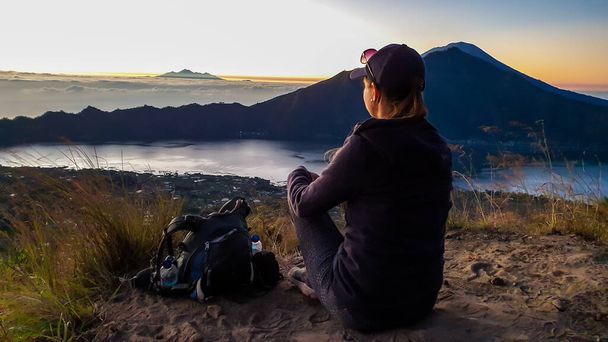 Eine Frau im Kapuzenpulli sitzt auf dem Gipfel des Mount Batur, Bali, Indonesien und wartet auf den Sonnenaufgang. Hinten liegt der Mount Rinjani (Lombok) und seitlich der Vulkan Agung. Nebel im Tal. Geheimnisvoll - Foto, Bild