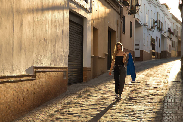 Mujer joven, hermosa y rubia, vistiendo una camiseta negra, chaqueta de piel azul en la mano y jeans, caminando por una calle solitaria y perdida durante la hora dorada. Concepto moda, belleza, puesta del sol. - Foto, imagen