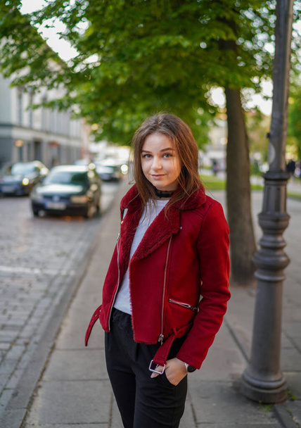 Beautiful Young Girl is Standing on the side Walk in Vilnius Old Town, Lithuania. Wearing Red jacket and Black Trousers. Beautiful Spring Day. Smiling. - Foto, imagen