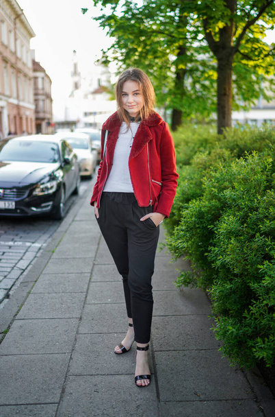 美しい少女はリトアニアのヴィリニュス旧市街の歩道に立っています。赤いジャケットと黒いズボンを着用。美しい春の日。笑顔. - 写真・画像