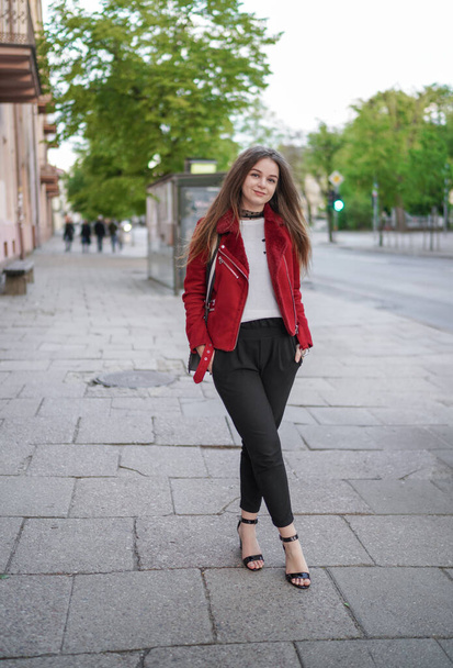 Beautiful Young Girl is Standing on the side Walk in Vilnius Old Town, Lithuania. Wearing Red jacket and Black Trousers. Beautiful Spring Day. Smiling. - Photo, image