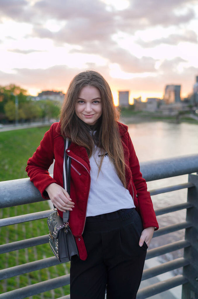 Beautiful Lithuanian Girl is Standing on the Mindaugas Bridge in Downtown of Vilnius, Capital of Lithuania. Wearing Red Jacket. Looking Back. Sunset Light - Photo, image