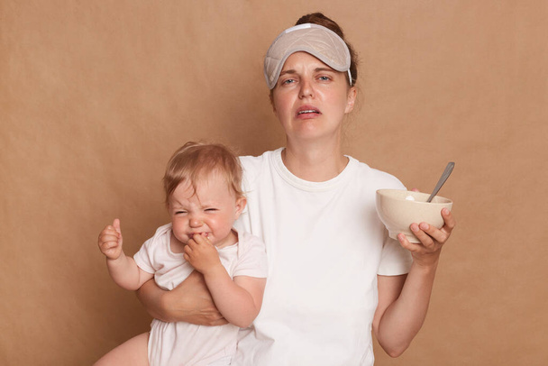 Portrait of crying sad stressed Caucasian woman wearing white T-shirt and sleeping mask, standing isolated over brown background, holding baby daughter and plate with food, needs to feed kid. - Foto, Imagen