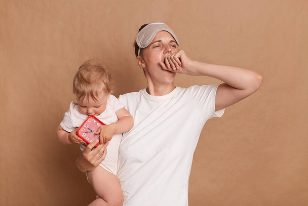 Sleepless woman wearing white t shirt and blindfold, yawning and covering her mouth, holding infant daughter, kid holds red alarm clock, has not enough sleep, posing isolated over brown background. - Foto, Imagem