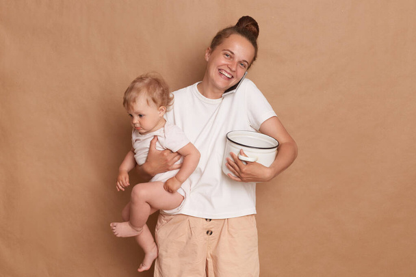 Plan intérieur d'une joyeuse mère blanche joyeuse avec une coiffure de chignon tenant sa fille en bas âge et un pot avec du porridge ou de la soupe, téléphone parlant isolé sur fond brun. - Photo, image
