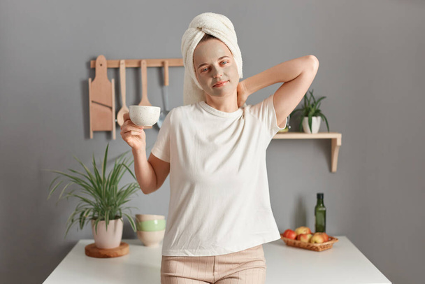 Indoor shot of young adult attractive woman with cleansing mask on her face standing in the kitchen at home, drinking her morning coffee, wakes up early. - Foto, afbeelding