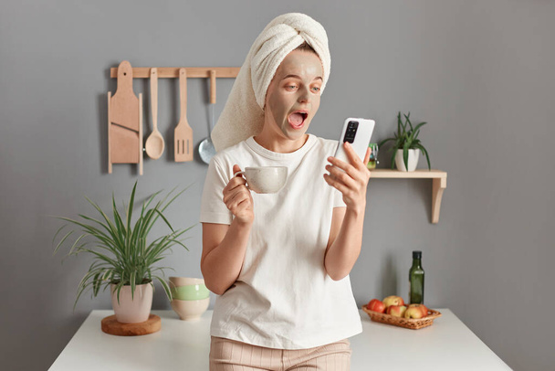 Indoor shot of shocked surprised woman wrapped in towel wearing white T-shirt standing in kitchen, drinking her morning coffee, doing cosmetic procedures, using mobile phone, surfing internet. - Foto, Imagen