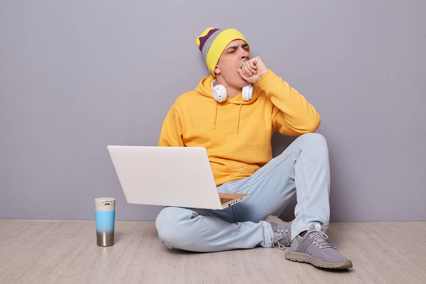 Horizontal shot of sleepy tired man wearing beanie hat, yellow hoodie and jeans posing against gray wall, being overworked, yawning, covering mouth with hand, working on notebook. - Fotó, kép