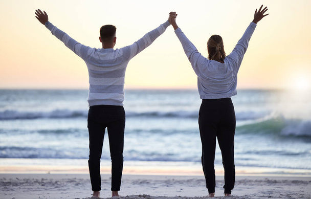 Couple at beach with hands up, freedom and travel, ocean waves and view, love and adventure together. Relationship, carefree and peace with man and woman with arms in air, back and sunrise in Bali. - Photo, Image