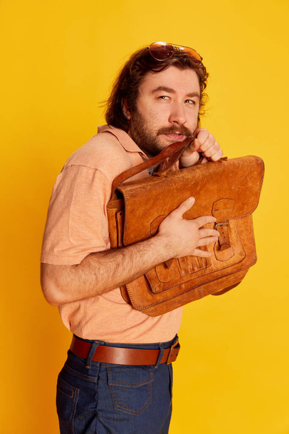 Fear. Portrait of bearded hairy man, shy scientist nerd in flared jeans standing with briefcase over yellow background. Retro fashion style, american culture, human emotions and ad - Photo, image