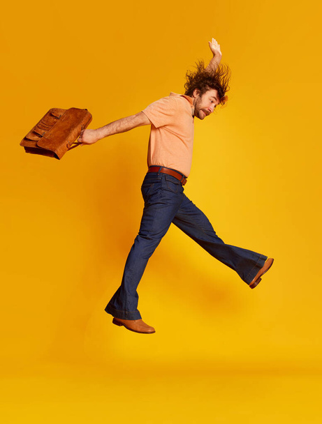 Excited mature bearded hairy man, hippie in stylish sunglasses and flared jeans running jumping over yellow background. Retro fashion style, american culture, emotions and ad - Photo, Image