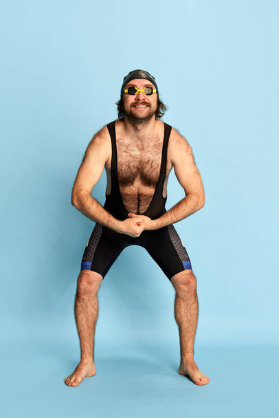 Comic portrait of funny bearded man in swimsuit and swimming cap and goggles having fun isolated over blue background. Concept of sport, active lifestyle and humor, funny meme emotions - Photo, Image