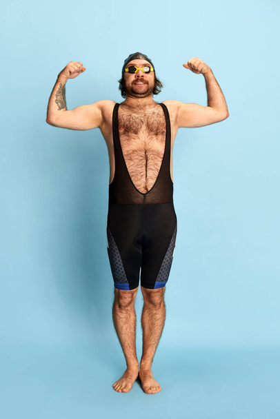 Comic portrait of funny bearded man in swimsuit and swimming cap and goggles having fun isolated over blue background. Concept of sport, active lifestyle and humor, funny meme emotions - Photo, image