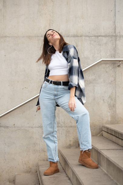 young caucasian girl with straight black hair, casual black and white checkered shirt and jeans, dancing alone in the street with headphones and cell phone. enjoying listening to music, looking at the sky. - Photo, image
