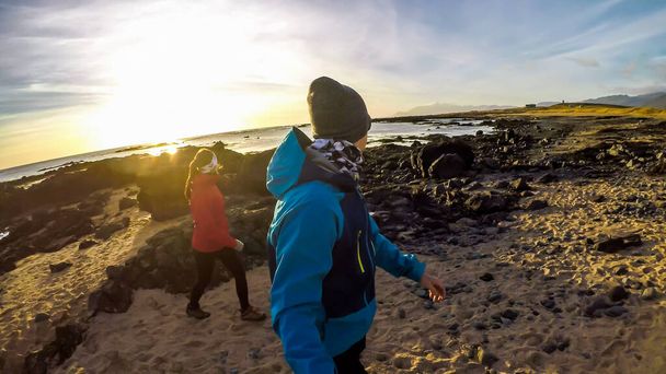 Una pareja caminando en la orilla del mar y observando focas jugando en el agua. El sol comienza a ponerse, extendiendo sus rayos a través del cielo. El cielo está lleno de nubes sedosas. Muchas rocas y piedras en la playa - Foto, imagen
