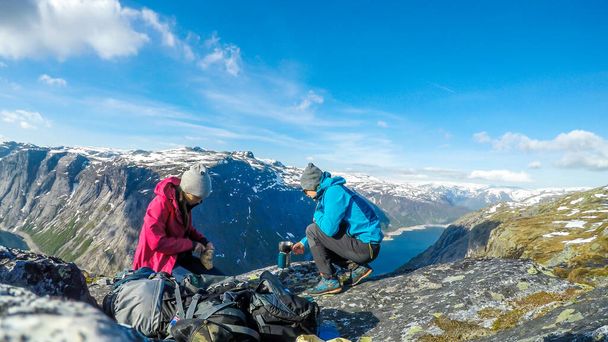 A couple having breakfast in the tall mountains with a view on a fjord-like lake. Girl is cutting the bread and the boy boils the water. Clear and sunny weather. Camping in the wilderness. - Foto, Bild