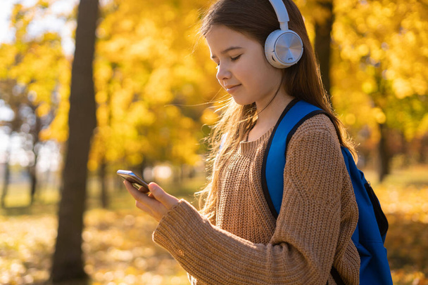 Happy schoolgirl relax in autumn nature. Pupil girl wear headphones in autumn park. Modern school education. School child listen music in fall forest. Enjoy elearning in headset. Copy space - Foto, afbeelding