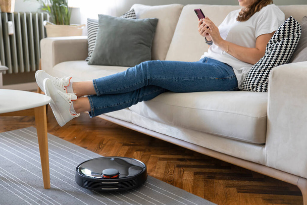 Robot vacuum cleaner cleaning the living room. Young woman enjoy rest, sitting on sofa at home - Fotó, kép