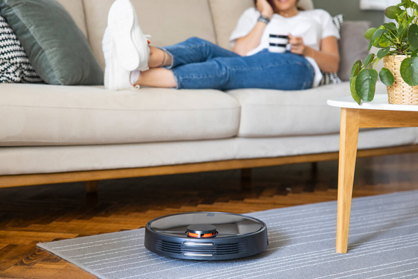 Robot vacuum cleaner cleaning the living room. Young woman enjoy rest, sitting on sofa at home - Fotó, kép