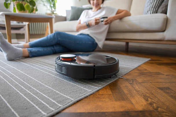 Robot vacuum cleaner cleaning the living room. Young woman enjoy rest, sitting on sofa at home - Foto, Bild