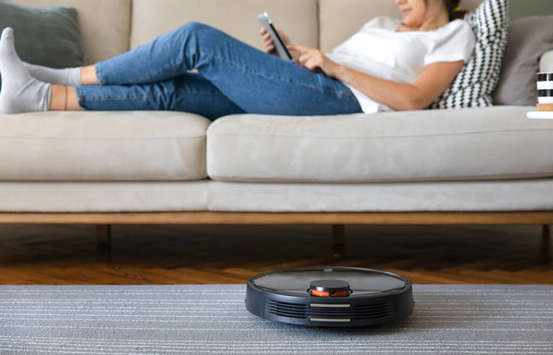 Robot vacuum cleaner cleaning the living room. Young woman enjoy rest, sitting on sofa at home - Фото, зображення