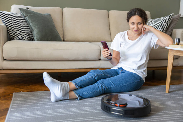 Robot vacuum cleaner cleaning the living room. Young woman enjoy rest, sitting on sofa at home - Photo, Image