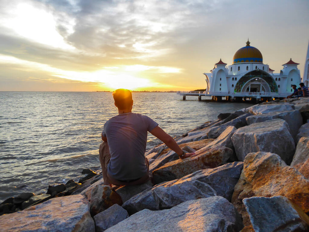 Ένας άνδρας που κάθεται μπροστά από το τζαμί Malacca Strait, Μαλαισία. Είναι Μνημείο Παγκόσμιας Κληρονομιάς. Προσεκτική σύλληψη κατά τη διάρκεια του ηλιοβασιλέματος, ο ήλιος δύει στη θάλασσα. Μοναδικός ταξιδιώτης. - Φωτογραφία, εικόνα