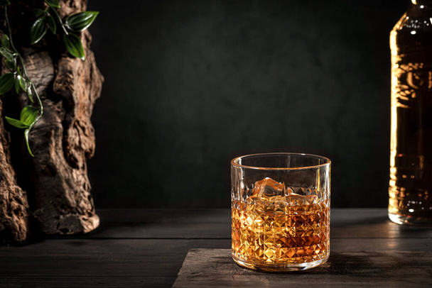 Vaso de whisky con hielo y botella sobre mesa de madera sobre fondo oscuro con espacio para copiar - Foto, imagen