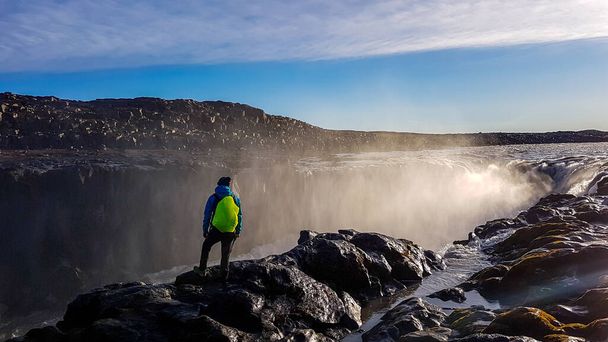A young man standing on top of a waterfall. He is wearing a blue jacket and  neon yellow backpack. Mist from the waterfall spreads in every direction.Water flowing in between the rocks. Wast waterfall - Foto, Bild